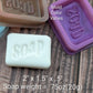 Bar of soap with 'soap' text, silicone mold, and text indicating mold color variation on a wooden surface.